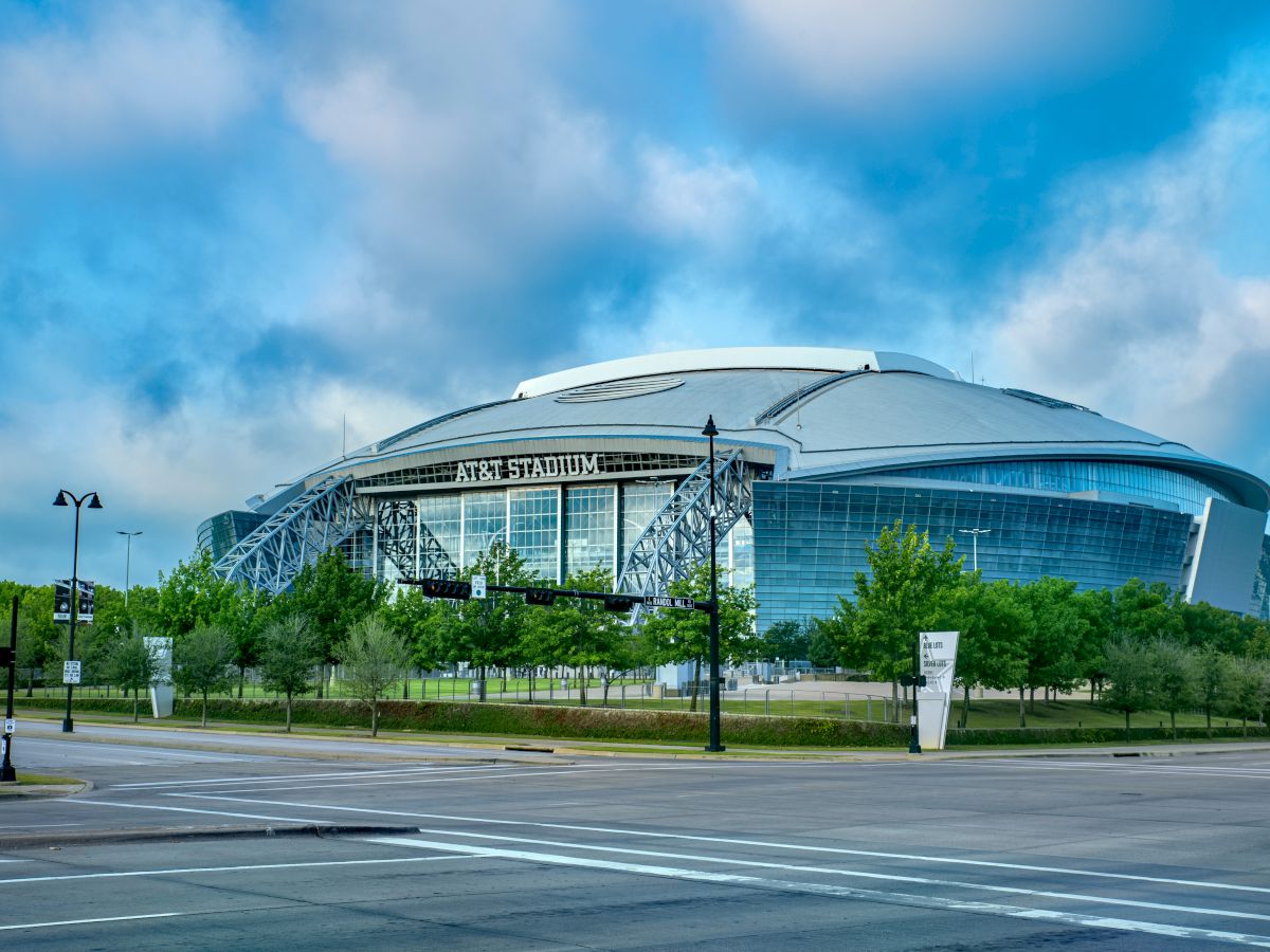 The image shows a large stadium surrounded by greenery, with a clear blue sky and scattered clouds in the background.