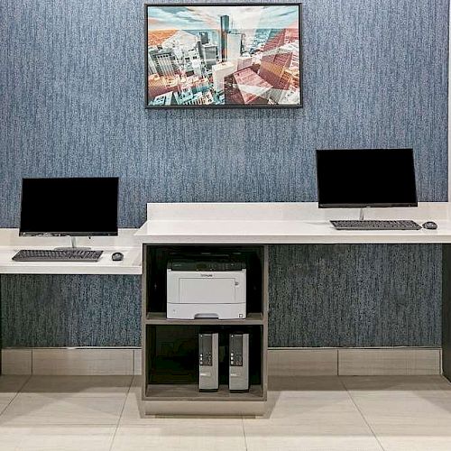 The image shows a small office space with two workstations, each equipped with a computer and keyboard, and a printer in the center.