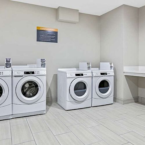 The image shows a laundry room with four front-loading washing machines and a countertop on the right, set against a light-colored tiled floor and walls.