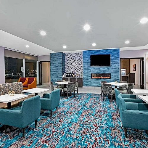 This image shows a modern lounge area with blue chairs, white tables, vibrant carpeting, and a TV mounted on the wall above a fireplace.