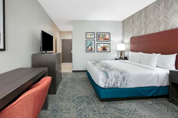 The image shows a modern hotel room with a large bed, TV, desk with a chair, bedside tables, and artwork on the walls.