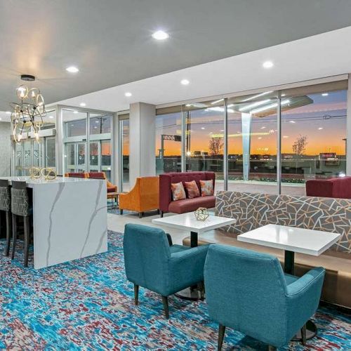 This image shows a modern, brightly lit lounge area with a mix of seating options, including blue and orange chairs and a patterned carpet, against a sunset backdrop.