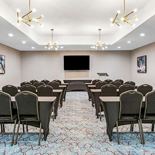 A small conference room with rows of chairs facing a podium and a large screen, modern ceiling lights, and framed artwork on the walls.