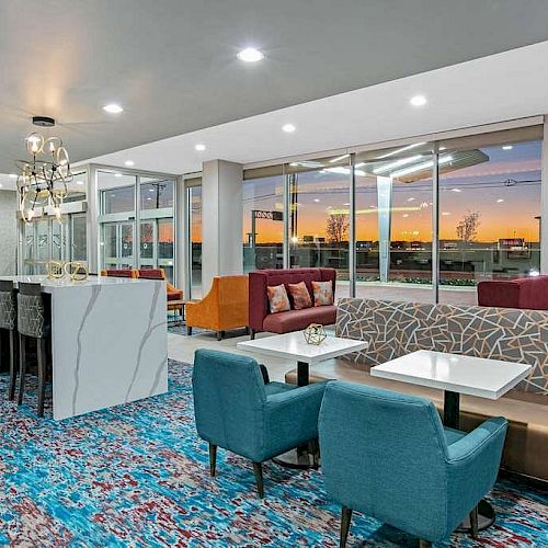 A brightly lit modern lounge area with colorful seating, marble tables, and large windows showcasing a sunset view.