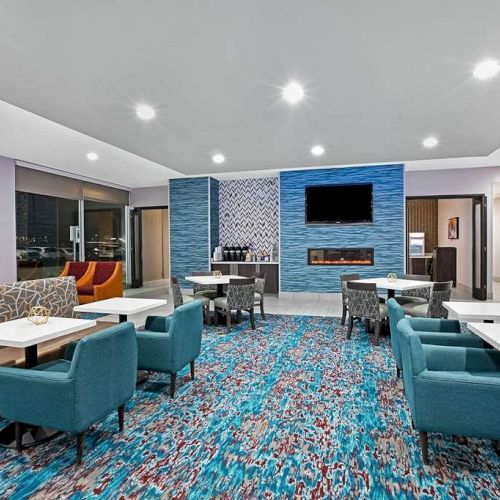 The image shows a modern lounge area with blue chairs, white tables, a patterned carpet, and a wall-mounted TV above a fireplace on a blue accent wall.
